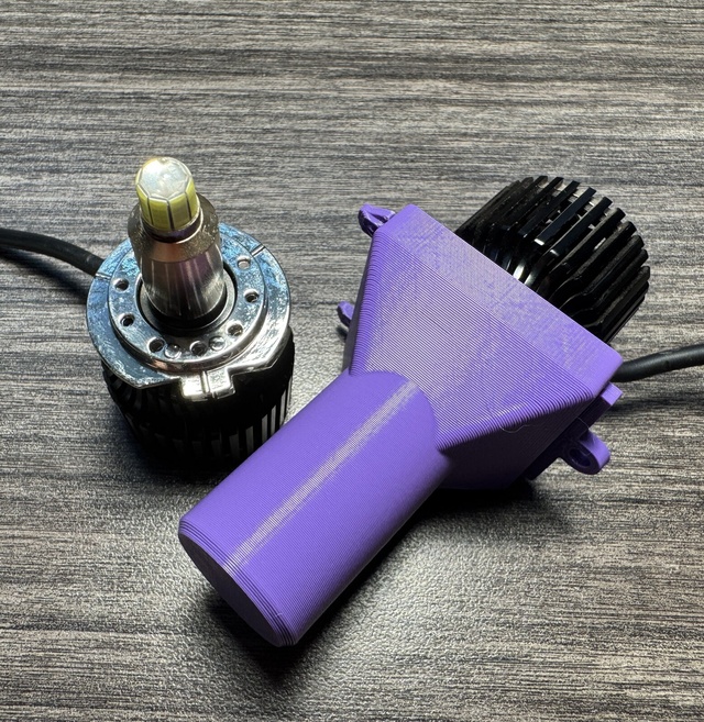 A purple 3D-printed cover with a long round tube and a square collar, lying next to an LED H7 headlamp bulb with a finned black heat sink